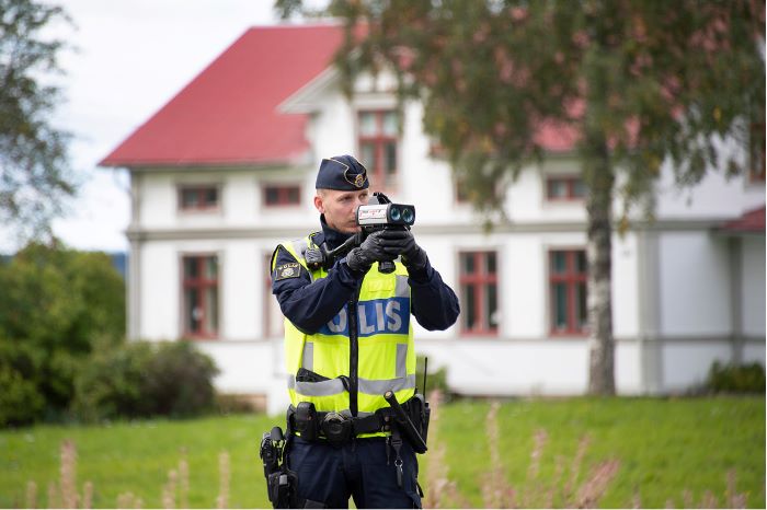 Polis som mäter hastighet med ett lantligt beläget hus i bakgrunden.