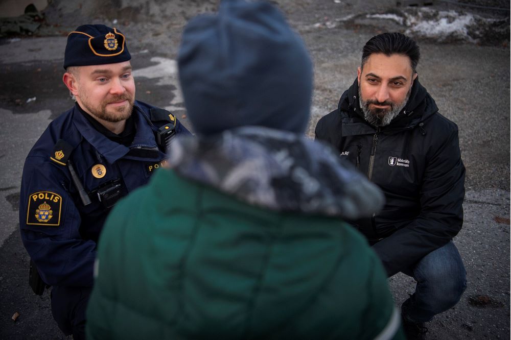 Fotografi av en polis i uniform och en annan vuxen som pratar med en ung människa som har ryggen mot kameran. Utomhus.