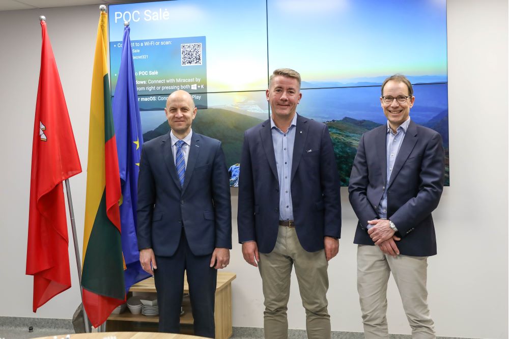 Three middle-aged men in blazers are standing in front of a large picture of green hills and blue clouds.